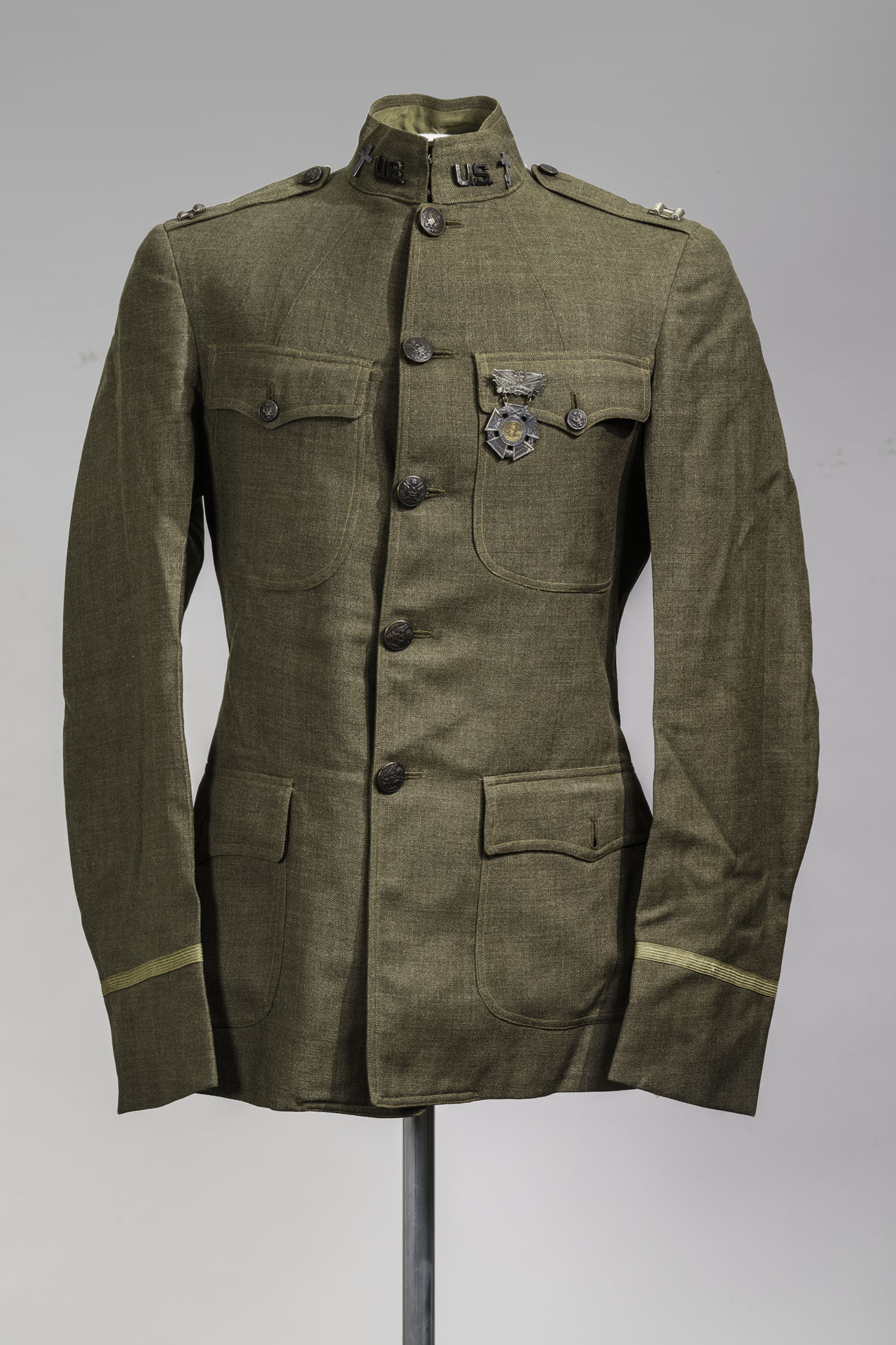 Modern photograph of an olive-green military tunic that buttons up the front and has four pockets, displayed on a dress form.