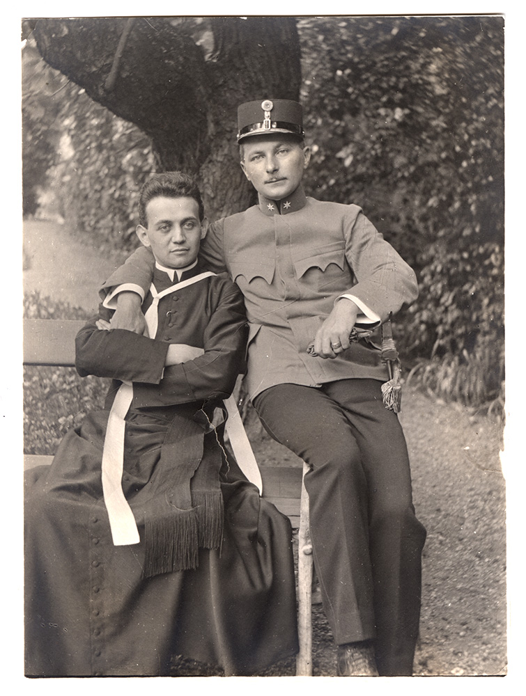 Black and white photograph of two white men sitting casually on a wooden bench in a park or garden. One man is cleanshaven, wearing dark clerical robes, a collar and other religious signifiers. The other man (dressed in a simple military tunic, pants and uniform hat, with a pencil mustache) has his arm slung around the shoulders of the man in robes.