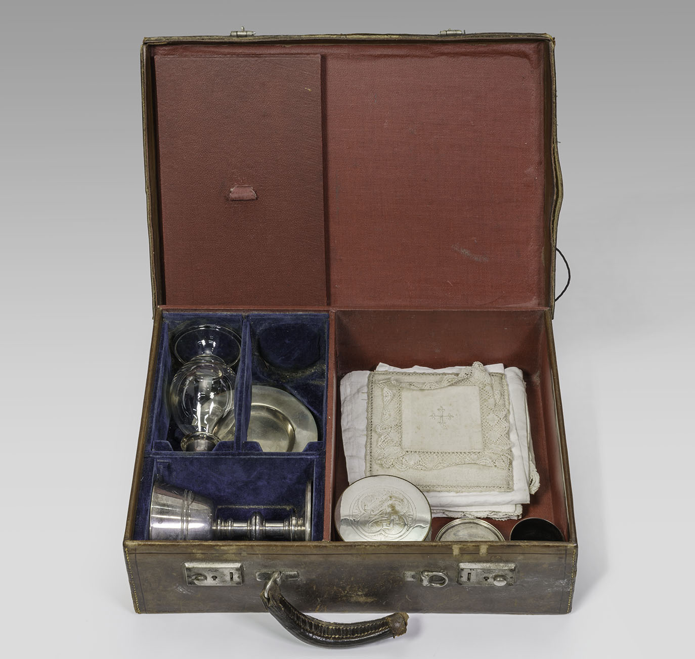 Modern photograph of a brown leather case that's been opened to show several compartments inside containing folded white loth, a silver-colored plate and chalice, and various metal and glass containers.