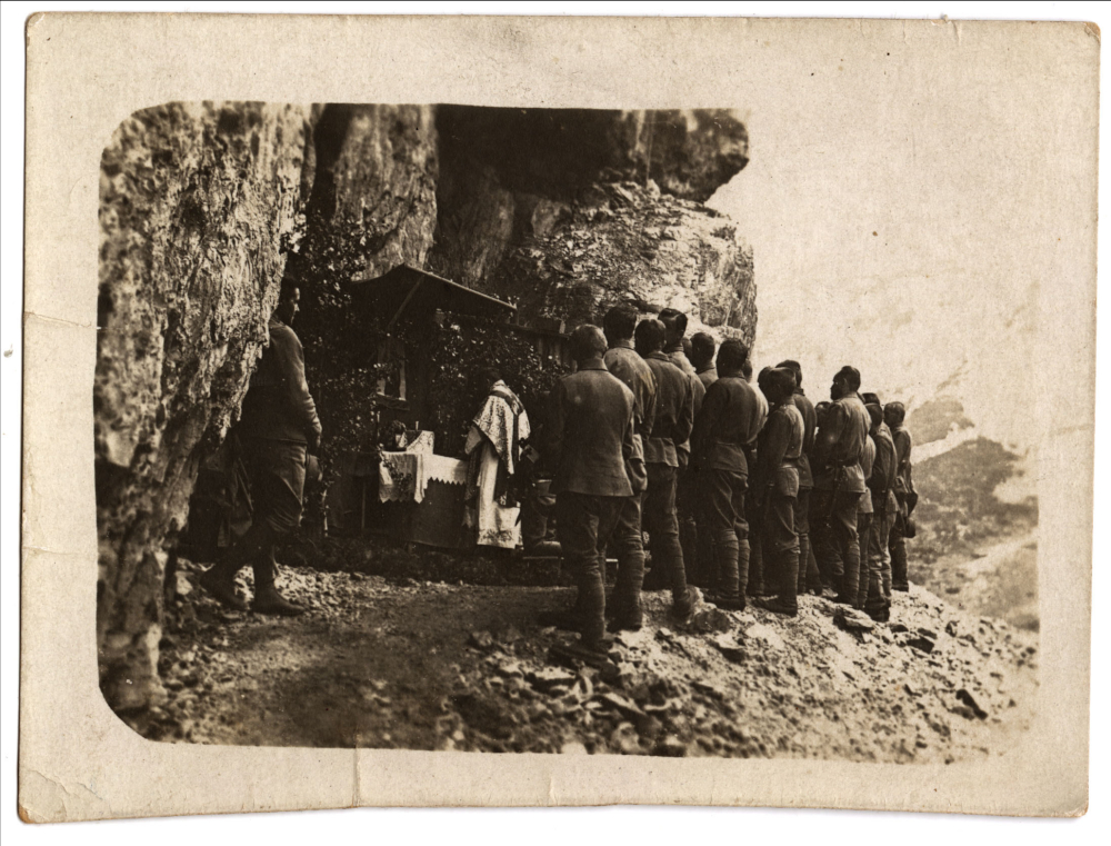Black and white photograph of a rocky mountainside with a group of soldiers in uniform standing and watching a man in Catholic priest robes doing something at a small Catholic altar covered in white cloth