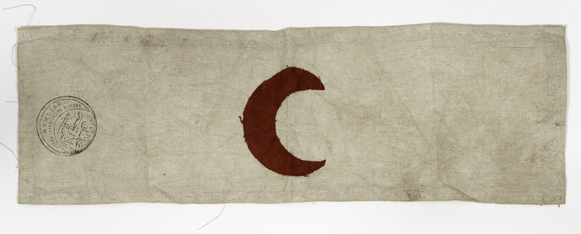 Modern photograph of an armband made of worn and aged cream-colored cloth with a red crescent shape embroidered in the middle