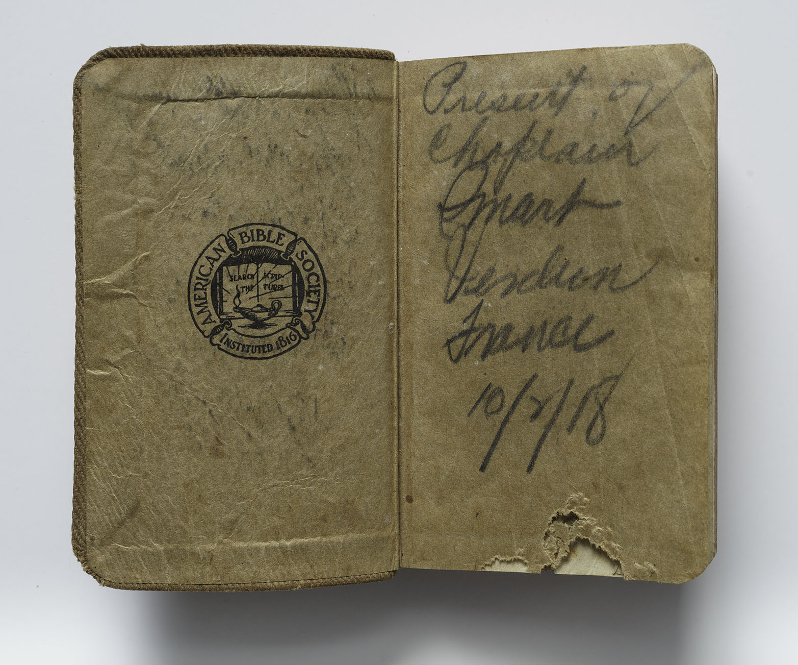 Modern photograph of an aged and torn book opened to the first page. The inside of the front cover is stamped with a crest for 'American Bible Society / Instituted 1816' depicting an ancient oil lamp in front of a book upon which is written, 'Read The Scripture.' On the front page is written in loose cursive 'Present of Chaplain Smart, Verdun, France, 10/8/18'
