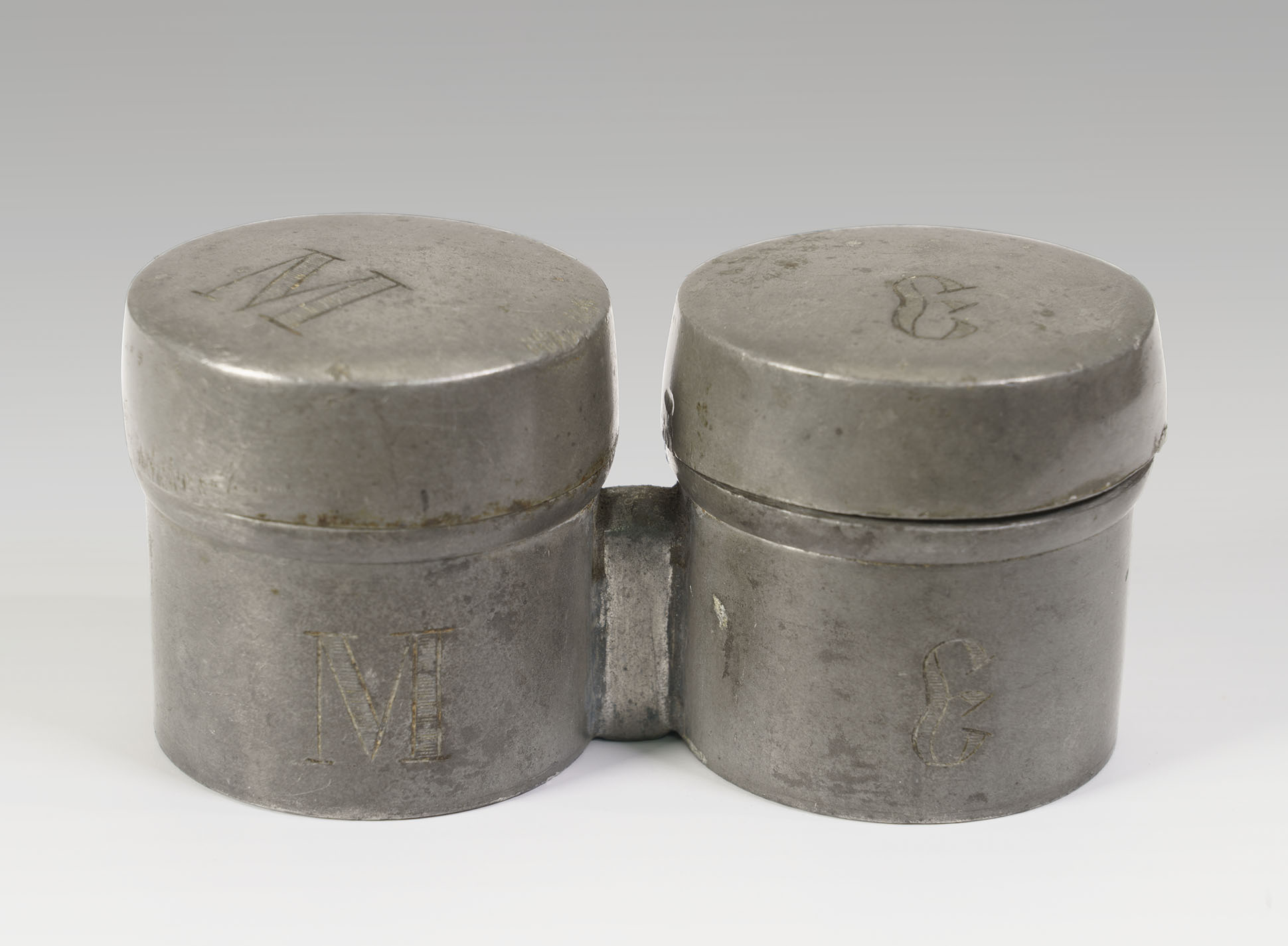 Modern photograph of two dull silver-colored round containers soldered together. Cyrillic letters are engraved on the containers and their lids.