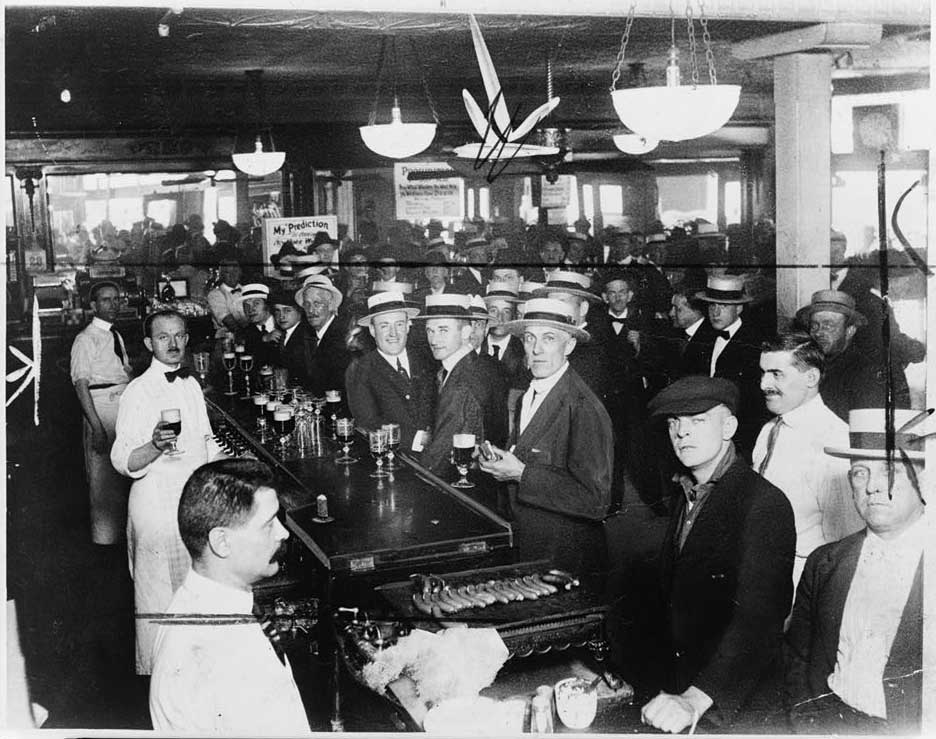 Black and white photograph of a crowded bar full of customers in suits and straw boater hats.