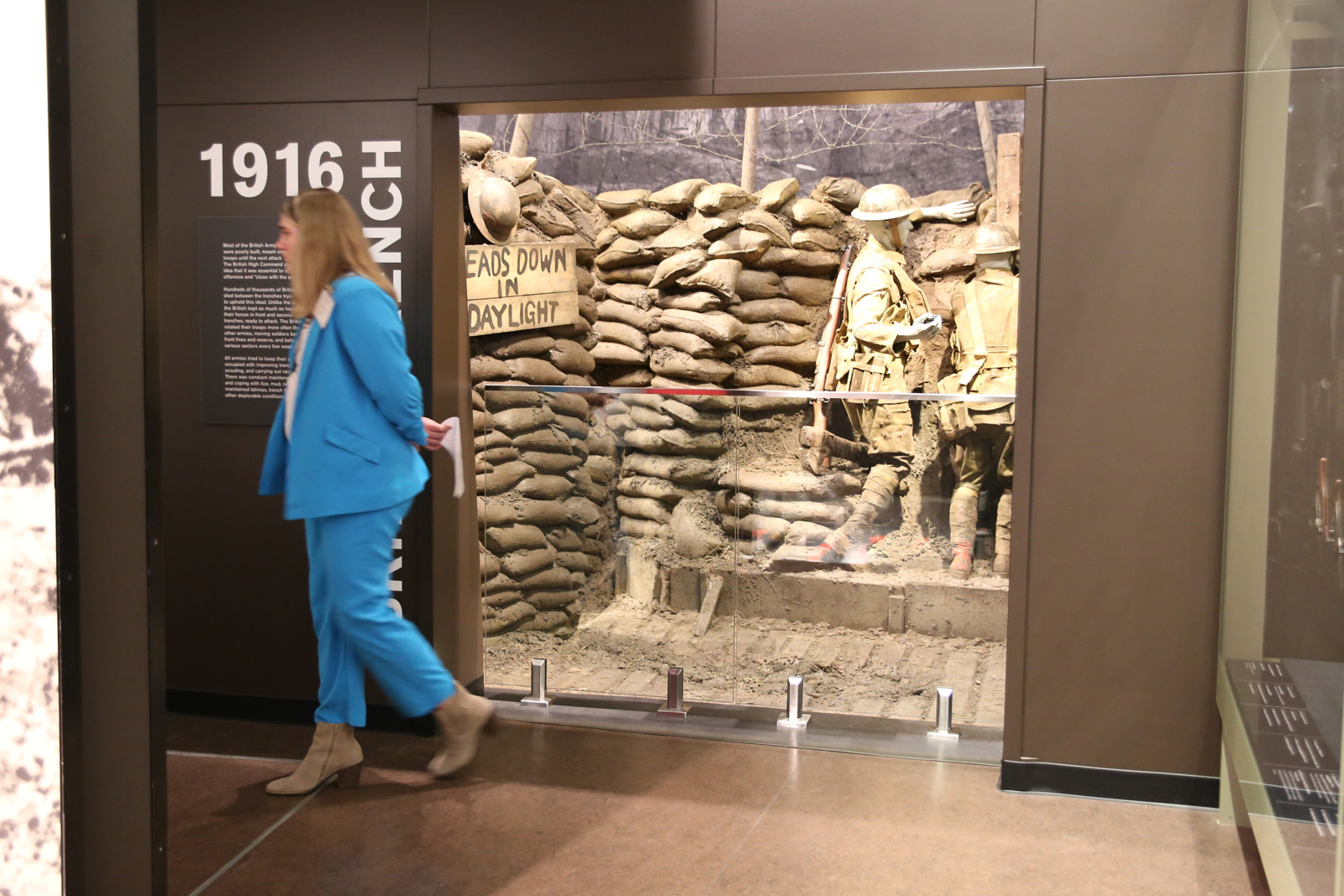 Modern photograph of a large opening in a museum exhibit that leads to a life-size cutaway model of a WWI trench. A young white person wearing a blue suit is walking away from the exhibit.