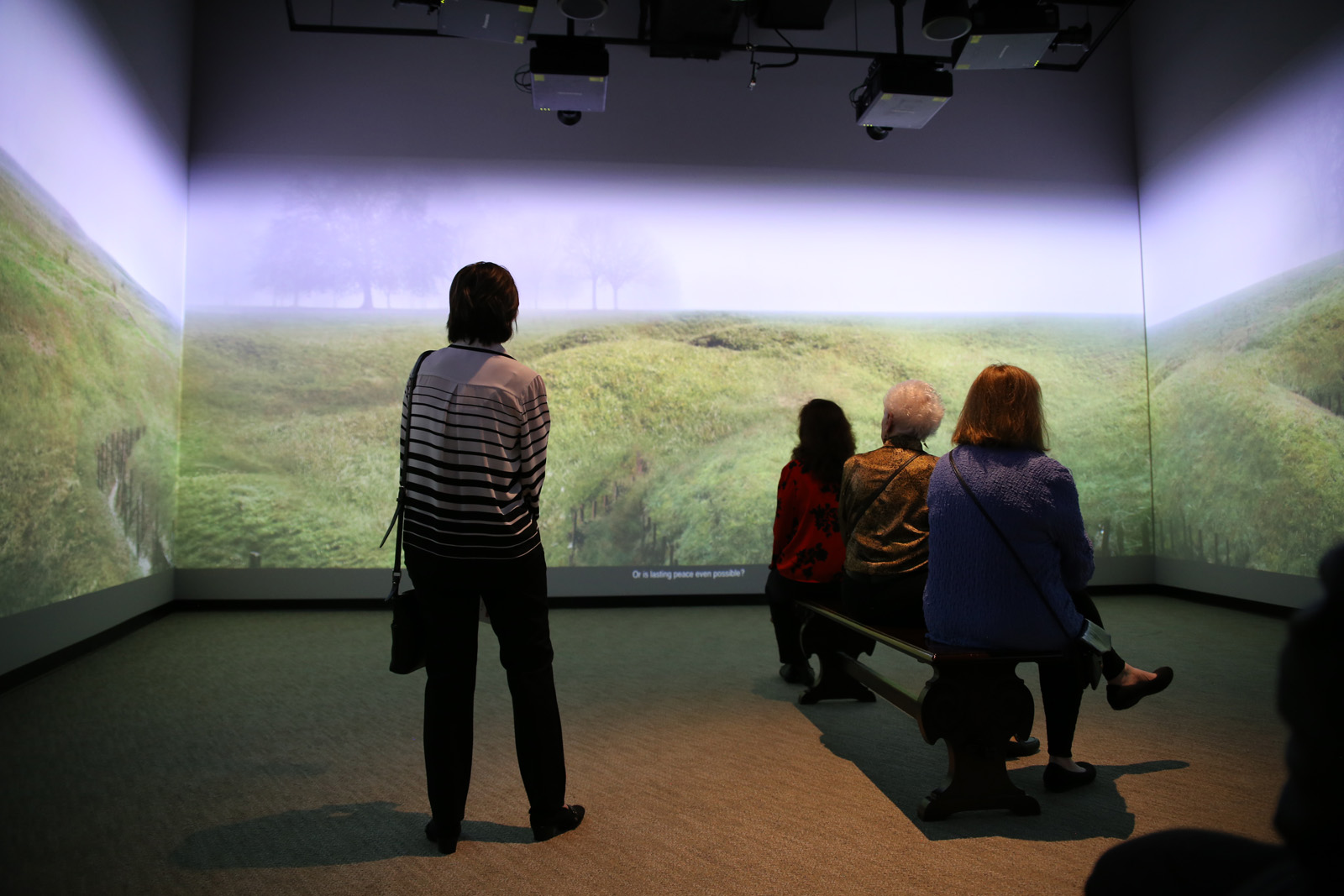 Modern photograph of four people sitting or standing in the center of a room, facing away from the viewer, while a video is being projected onto three walls around them - currently a scene of a wide green field or grassland.