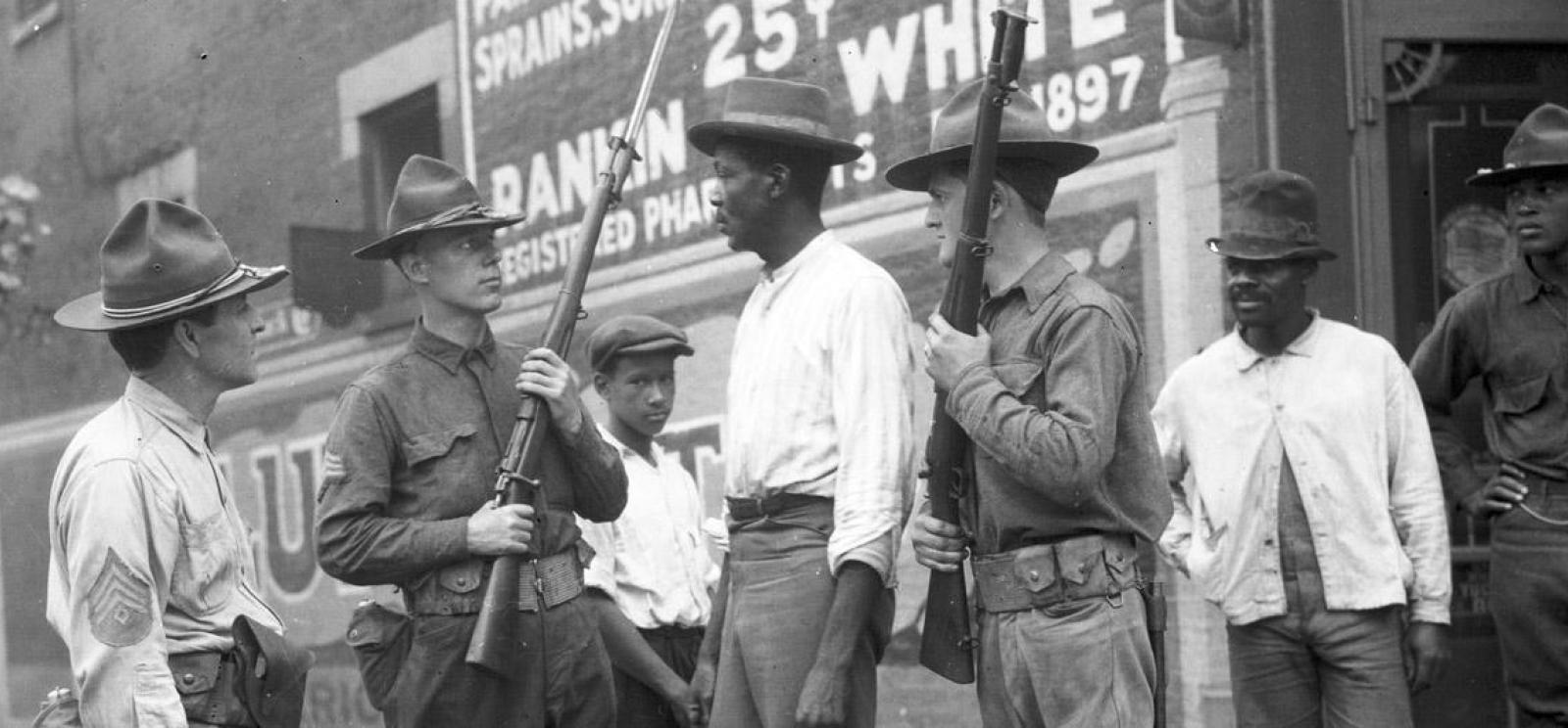 Black and white photograph of three white men in National Guard uniform holding rifles surrounding a Black man in civilian clothing. Two Black men in civilian clothing observe off to the side. A Black man in military uniform also observes.