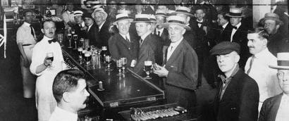 Black and white photograph of a crowded bar with bow-tied bartenders and customers wearing suits and straw boater hats.