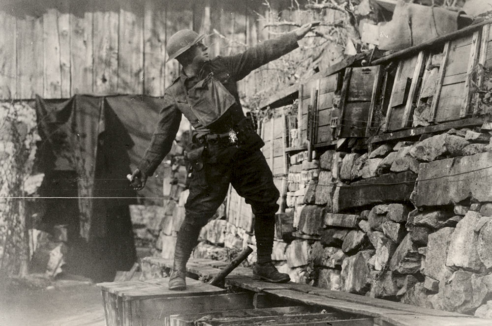 Black and white photograph of a white man in military combat gear in a trench. He is leaning back on his right foot, left arm extended out and right arm extended down, about to throw the grenade in his hand in an overhead throw.