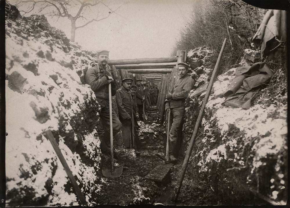 Black and white photograph of a snowy trench stretching directly away from the viewer. It is lined with wooden supports and wooden roof scaffolding. Several soldiers holding shovels pose for the photograph in the trench.