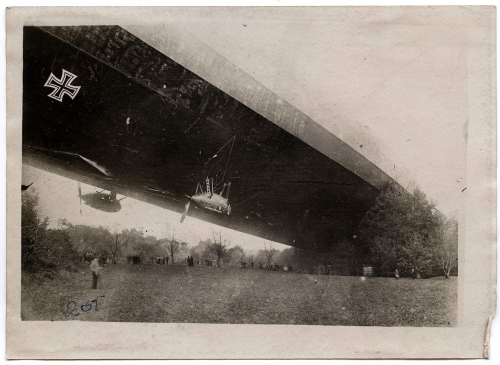 Black and white photograph. A long rigid-bodied airship stretches across the frame with one end resting in a stand of trees.