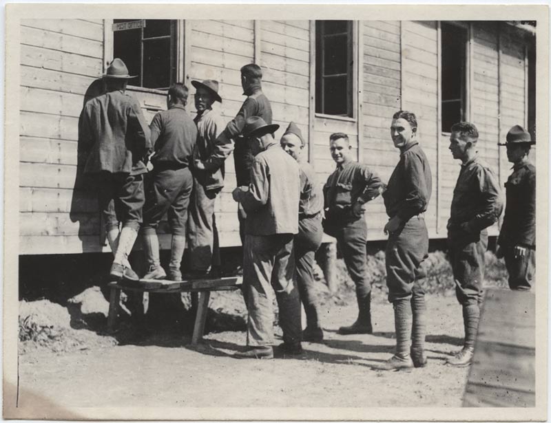 Black and white photograph of a messy line of men dressed in informal military uniform. They are standing in front of an open window of a wooden building. Some of them have turned their heads and are smiling at the cameraperson.