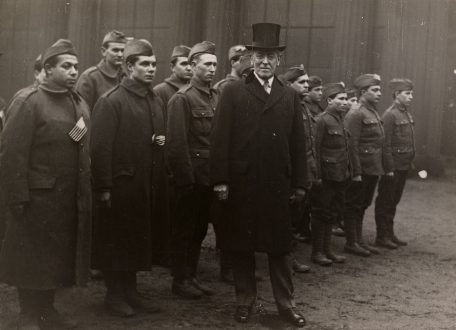 Black and white photo of Woodrow Wilson in a top hat and overcoat standing in front of a small group of lined-up soldiers in military uniform.