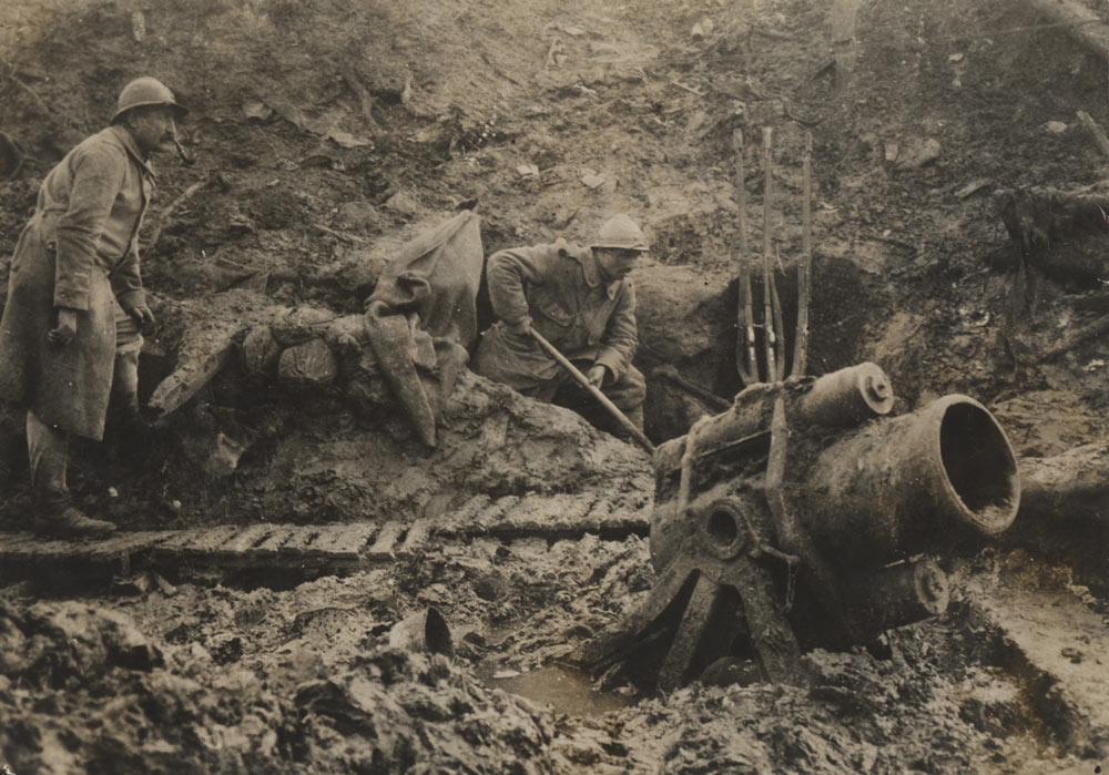 Sephia photograph of two soldiers in a muddy trench operating a short stubby trench gun.