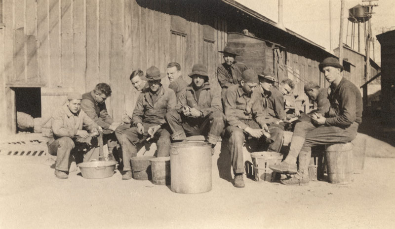 Black and white photograph of a dozen men seated on some sort of concrete structure with buckets and barrels in front of them. They are peeling potatoes with knives.