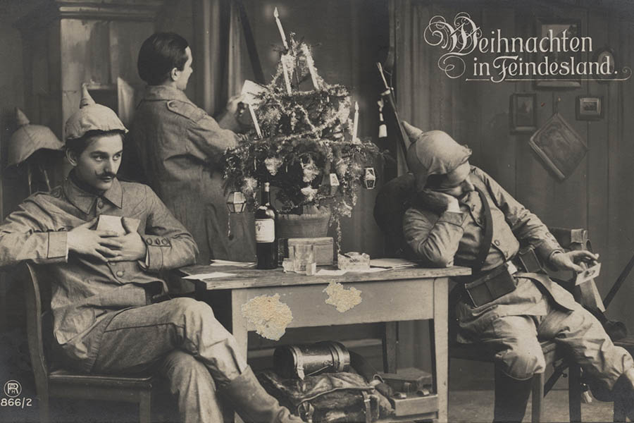 Black and white photograph of three German soldiers. Two are seated on either side of a small table with a small Christmas tree on it. The third man is standing by the table looking at a sheet of paper.