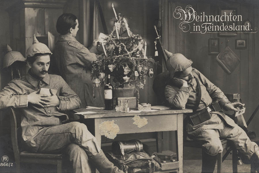 Photographie en noir et blanc de trois soldats allemands. Deux sont assis de chaque côté d'une petite table avec un petit sapin de Noël dessus. Le troisième homme est debout près de la table et regarde une feuille de papier.