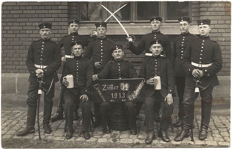 Black and white photograph of nine men in Bavarian military uniform seated and standing. Two of the soldiers are crossing their swords. One soldier is holding a sign. Two soldiers hold large mugs of beer.