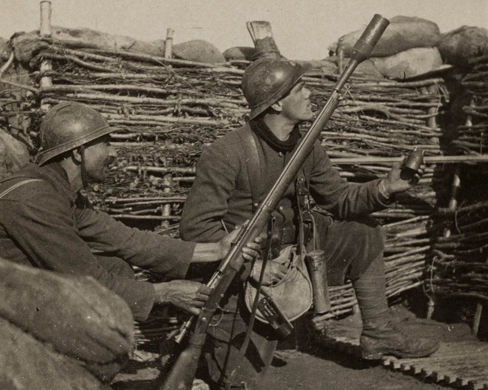 Black and white photograph of two French soldiers in combat gear, kneeling in a trench. One of them holds a rifle with a grenade launcher attached to the end. The other one holds a grenade.