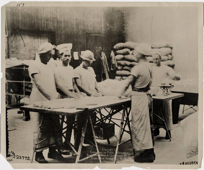 Five men gathered around a table covered in piles of bread dough. The men are either mixing the dough or kneading it.