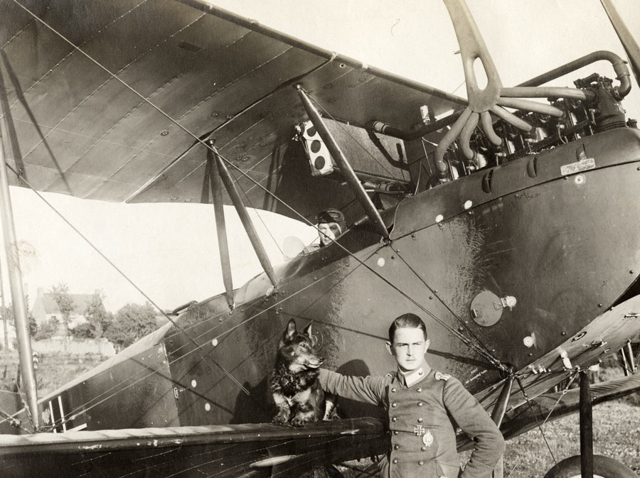 Black and white photograph of a WWI-era aircraft with a pilot in the seat. Another soldier stands on the ground in front of the wing, petting a dog that is seated on the wing.