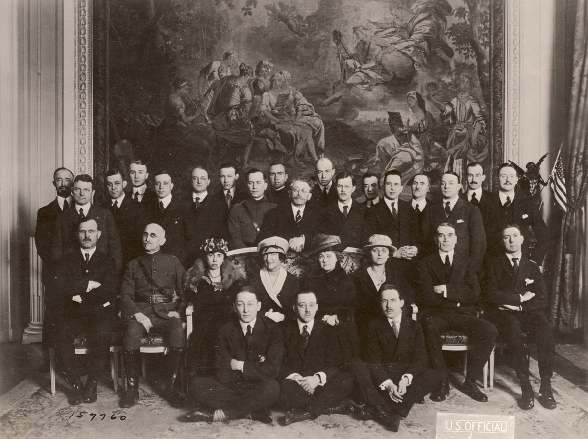 Photographie en noir et blanc d'un groupe d'hommes et de femmes en costumes et robes de tailleur posant sous un grand tableau