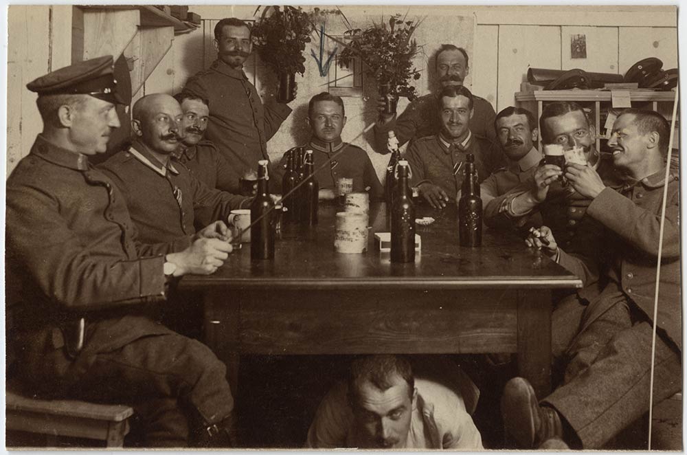 Black and white photo of a raucous group of German soldiers gathered around and under a table drinking bottles of beer. 