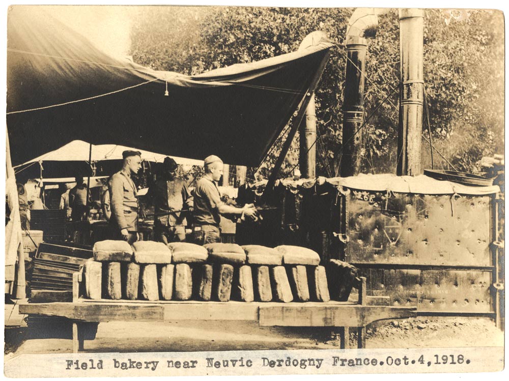 Black and White photograph of American WWI soldiers in uniform manning large metal outdoor ovens under a tent or tarp, with dozens loaves of bread stacked on end on a rack or bench in the foreground
