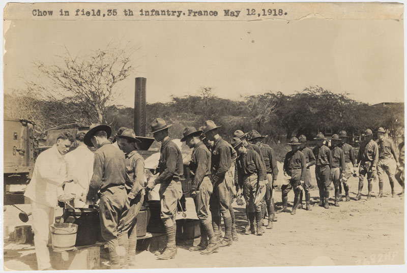 Photo sépia de plusieurs soldats servant de la nourriture à une longue file de soldats dans un champ ouvert.