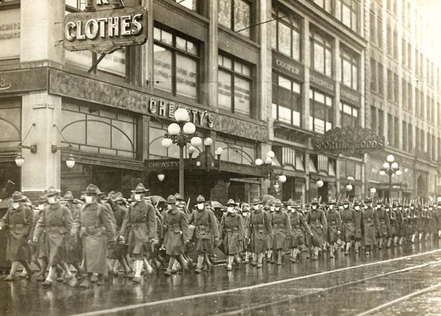 Black and white photograph of a regiment of soldiers marching down a city street. The soldiers are wearing white masks.