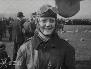 Animated gif of black and white film footage. A white woman in aviator hat, goggles and jacket looks at the camera and laughs.