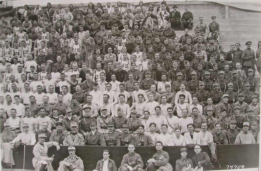 Black and white photograph of a section of the stands in a stadium. The people in the seats are dressed in the sports uniforms for various countries, grouped together by uniform.
