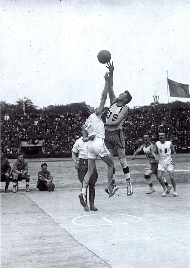 Black and white photograph of a basketball game. Two athletes from opposing teams leap into the air after the basketball.