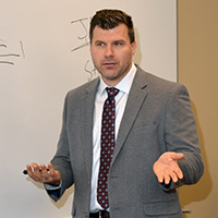 Headshot of a white man with short brown hair standing in front of a whiteboard