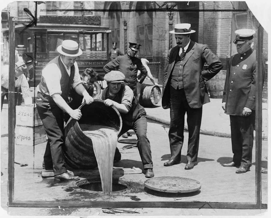 Black and white photograph of two men in shirtsleeves and vests pouring a large barrel of alcohol down an open sewer manhole. Three more men stand and observe.
