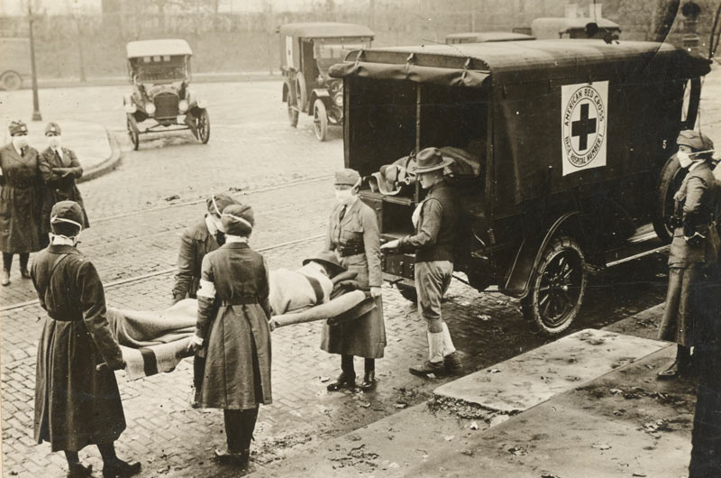 Black and white photograph of masked Red Cross orderlies carrying a stretcher with a prone person on it to an automobile with the Red Cross logo on it.