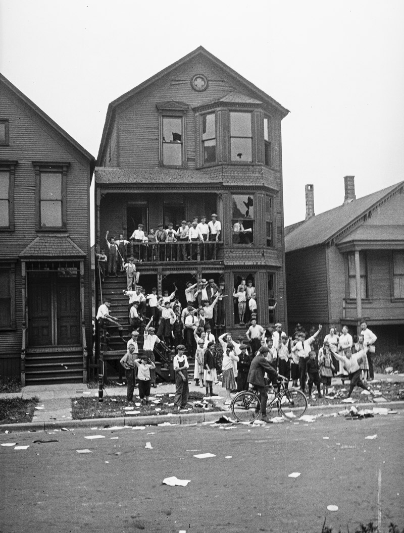Black and white photograph of a house across the street. The front yard and porch are full of white children. Papers and debris are scattered around the scene.