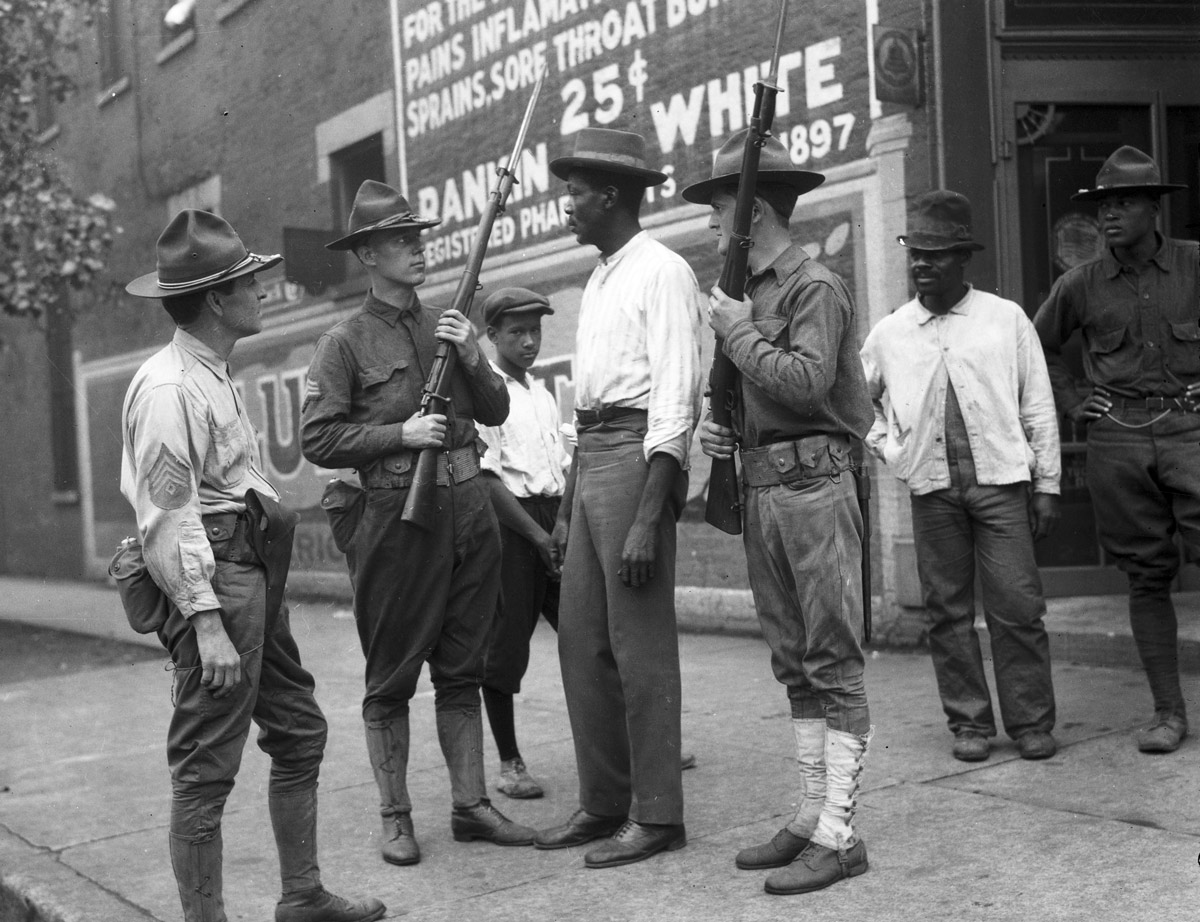 Black and white photograph of three white men in National Guard uniform holding rifles surrounding a Black man in civilian clothing. Two Black men in civilian clothing observe off to the side. A Black man in military uniform also observes.
