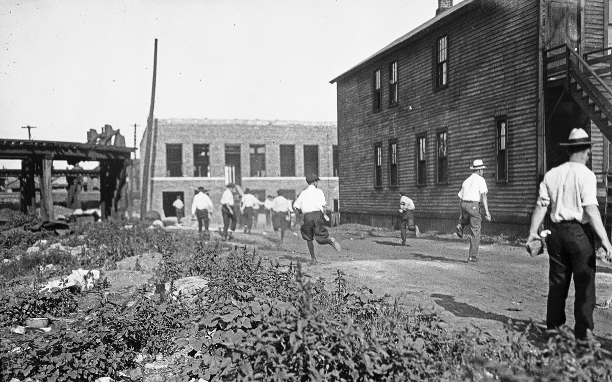 Black and white photograph of a street outside several apartment buildings. A group of white men run down the street away from the viewer. Some of them are in mid-throw.