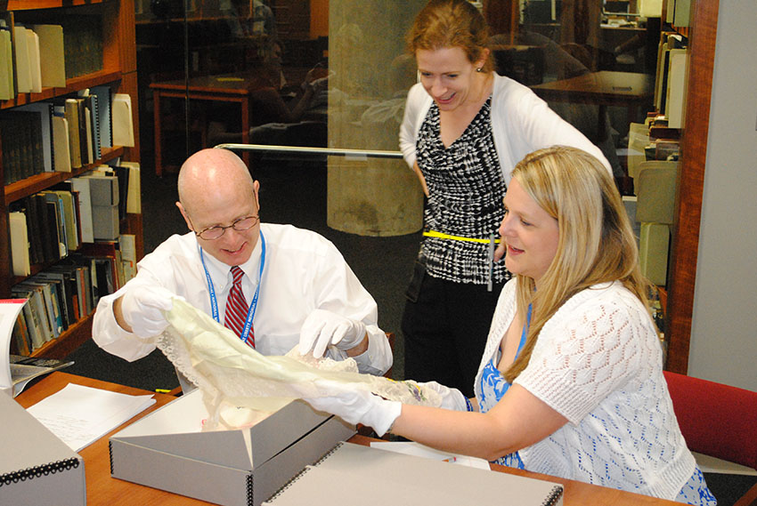 Photograph of 3 modern-day white adults dressed in business casual clothing in a research library. They are examining a textile artifact.