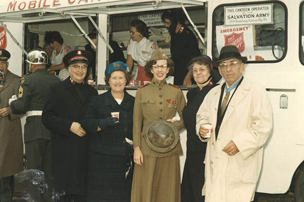 Vintage color photograph of an older white man in a trench coat and hat standing next to several people dressed in WWI-era clothing and uniforms, in front of a Salvation Army mobile canteen.