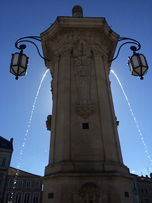 Modern photograph of a tall stone fountain silhouetted with the sun behind it.