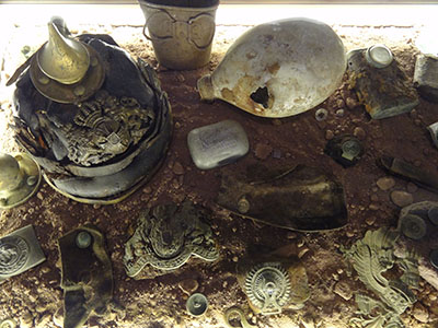 Modern photograph of various WWI-era artifacts including an engraved steel helmet, lying in the dirt.