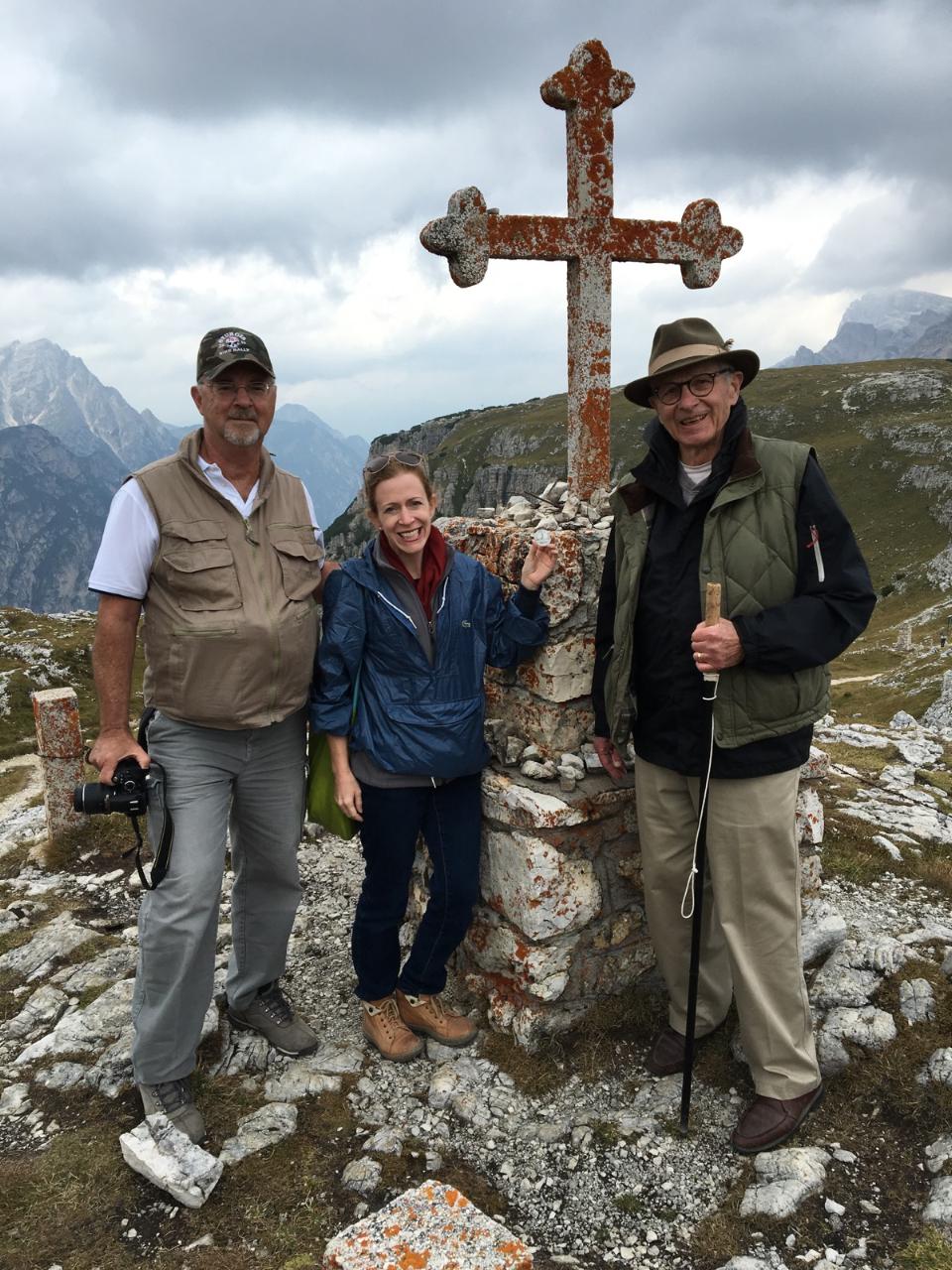 A white woman and two white men dressed in hiking gear posing in front of a cross memorial on a cairn.