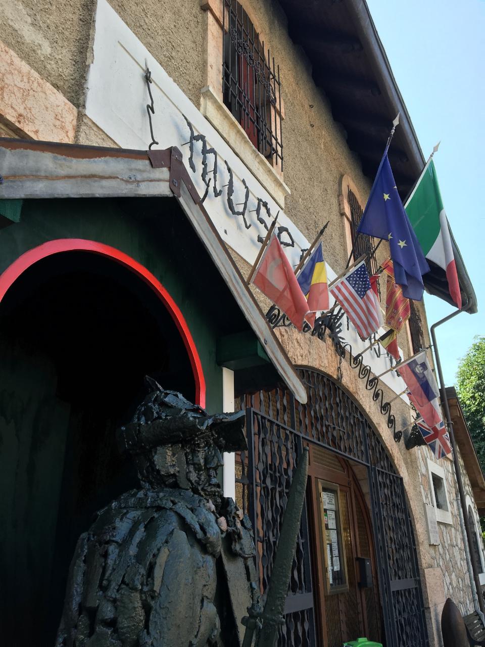 View of the front of a medieval building adorned with many flags. Standing close to the viewer is a sculpture of a soldier holding a sword.