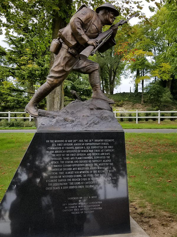 Photograph of a bronze statue on a plinth. The statue depicts a doughboy striding forward with his rifle in hand. The plinth is smooth black stone and engraved with text.