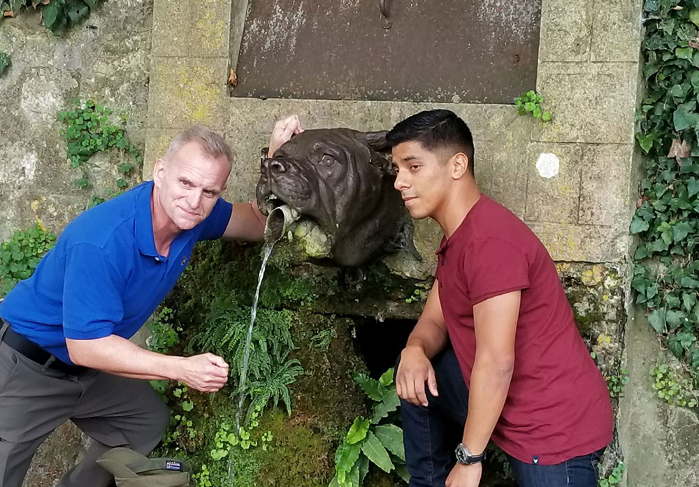 Two men pose in front of a fountain shaped like a dog's head.
