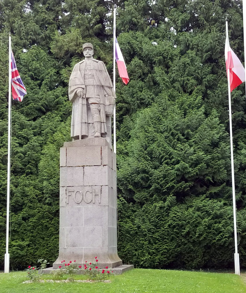 Photo of a statue on a plinth in front of green trees. The statue is of a man dressed in a greatcoat and military hat.