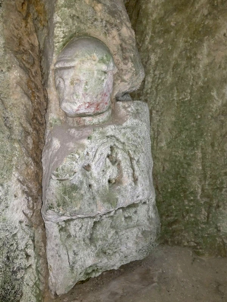 Photograph of a crudely carved figure wearing a steel helmet emerging from a cave wall.