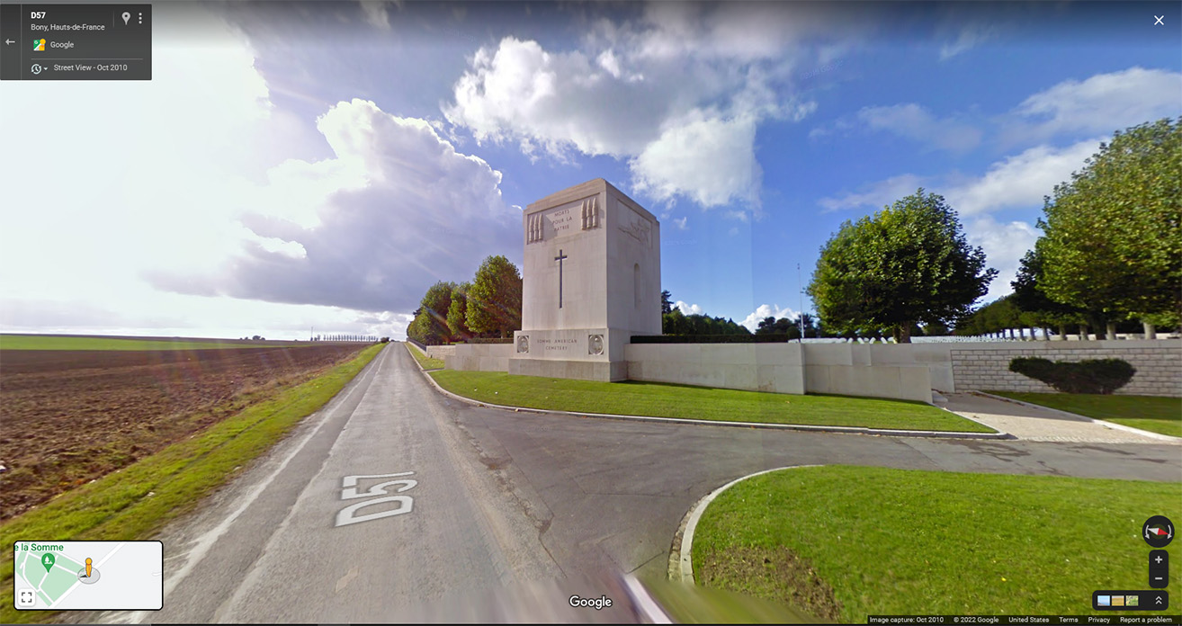 Google Street View of a road running past a large square stone monument at the entrance to a cemetery.